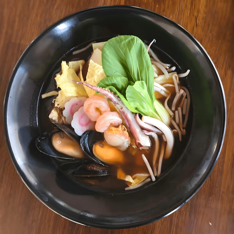 A black bowl of Koji Ramen seafood noodle soup with shrimp, mussels, squid, and bok choy.