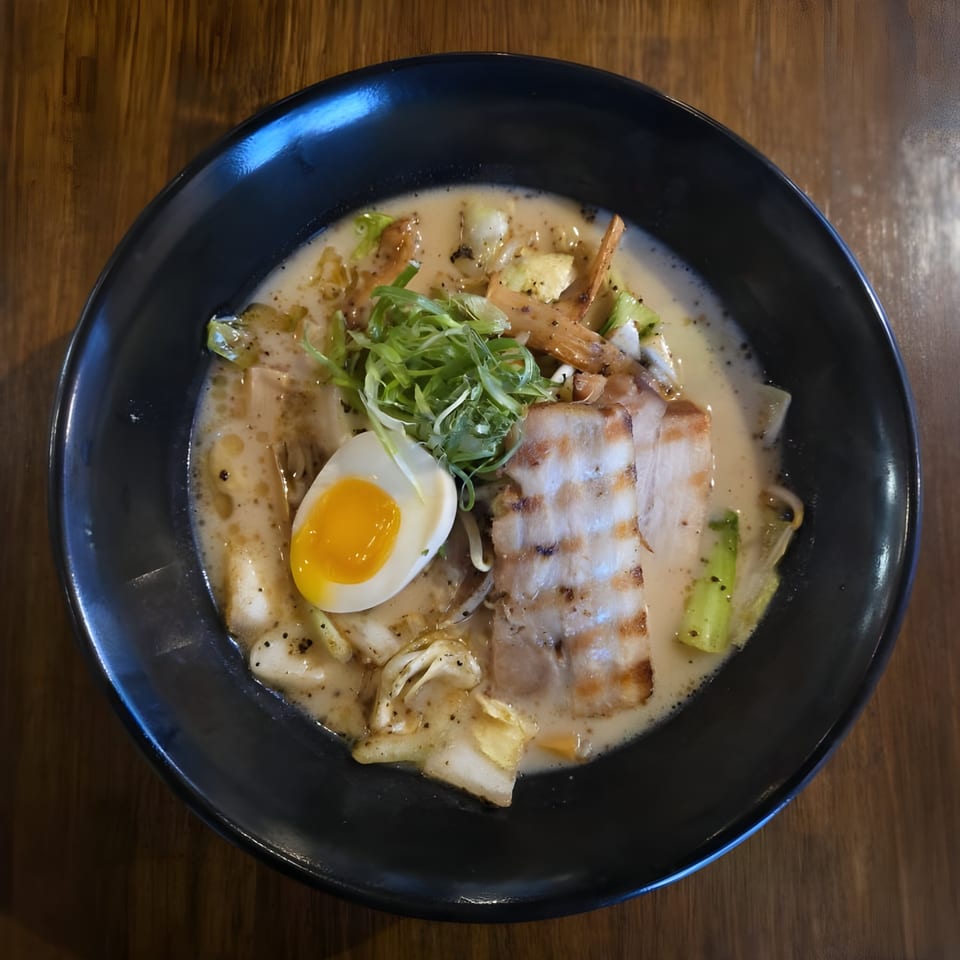 A black bowl of Koji Ramen with pork belly, soft-boiled egg, and green onions.