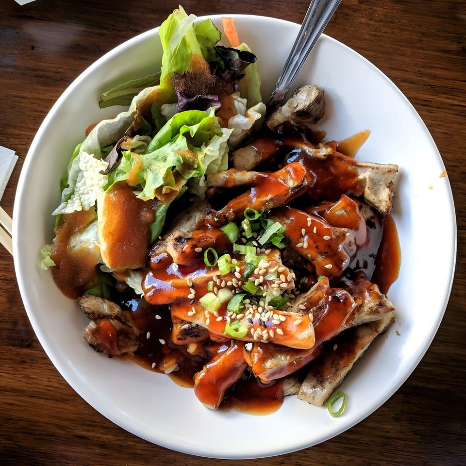 Teriyaki chicken with sesame seeds, green onions, and a side salad in a white bowl.